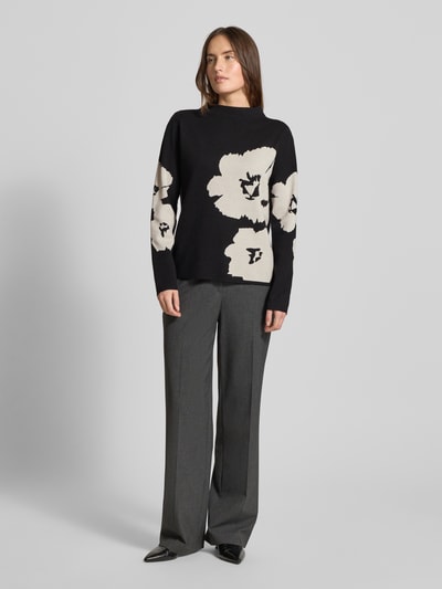 Frauenmodel trägt Strickpullover mit geripptem Stehkragen von Betty Barclay, Viskose-Mix, lockerer Schnitt, schwarze Grundfarbe, weiße Blumenmuster, kombiniert mit grauer weiter Hose, Ganzkörperansicht von vorne.
