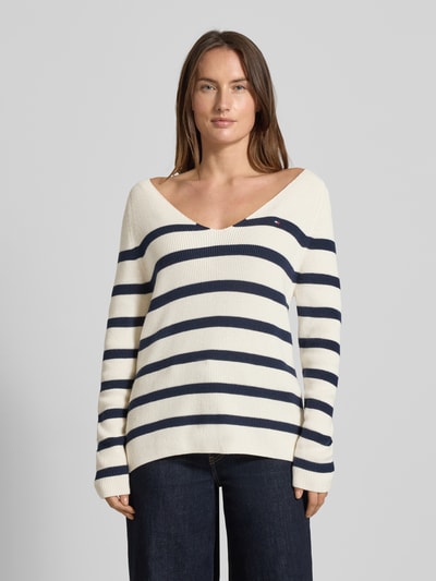 Frauenmodel trägt Regular Fit Strickpullover aus reiner Baumwolle von Tommy Hilfiger in Optik: Allover-Muster, gestreift, Rippstrick, Logo-Stitching, V-Ausschnitt, Langarm, Raglanärmel, Farbe Beige mit dunkelblauen Streifen; Ganzkörperansicht von vorne.