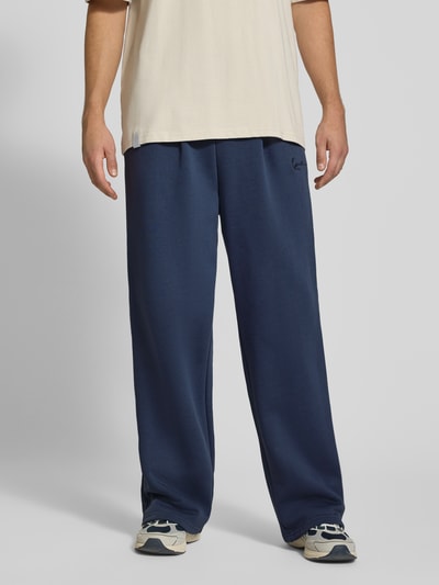 Männermodel trägt Sweatpants mit Logo-Stitching und Eingrifftaschen von Karl Kani in Dunkelblau, Baumwoll-Mix, Wide Fit, Ganzkörperansicht von vorne.