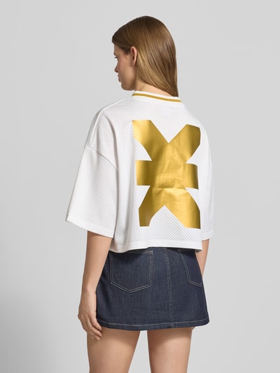 Frauenmodel trägt Cropped T-Shirt mit rückseitigem Logo-Print von Karo Kauer, Damen T-Shirt in Weiß mit goldfarbenem Logo-Print, Loose-Fit, Rundhalsausschnitt, überschnittene Schultern, Label-Print und -Stitching, Rückenlänge bei Größe S: 47 cm, Ganzkörperansicht Rückansicht.