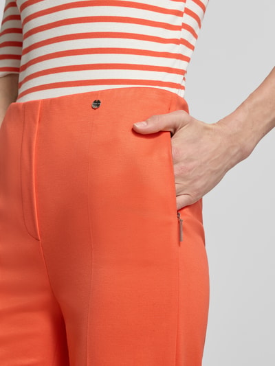 Mężczyzna model nosi Stoffhose aus Viskose-Mix mit Bügelfalte von Marc Cain w kolorze pomarańczowym, francuskie kieszenie, detal z logo, prosta nogawka, krój straight leg fit, luźny materiał z domieszką stretchu, szerokość w rozmiarze 36: 36 cm, długość wewnętrzna nogawki w rozmiarze 36: 80 cm, zapięcie na haczyk i zamek, zbliżenie na górną część.