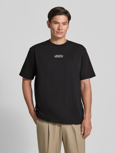Männermodel trägt Regular Fit T-Shirt aus reiner Baumwolle Modell 'Oliver' von Wood Wood, Schwarz, Logo-Stitching, Ganzkörperansicht von vorne. Rückenlänge bei Größe M: 73 cm.
