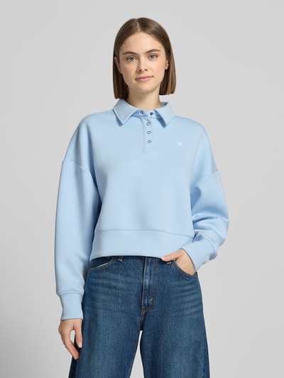 Frauenmodel trägt Cropped Pullover mit Viskose-Anteil von Guess Jeans in Hellblau mit Polo-Kragen und Logo-Stitching, Nahaufnahme Oberkörper.