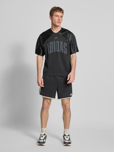 Männermodel trägt T-Shirt mit V-Ausschnitt und Logo-Print von adidas Sportswear in Schwarz, lockerer Schnitt, Ganzkörperansicht von vorne.