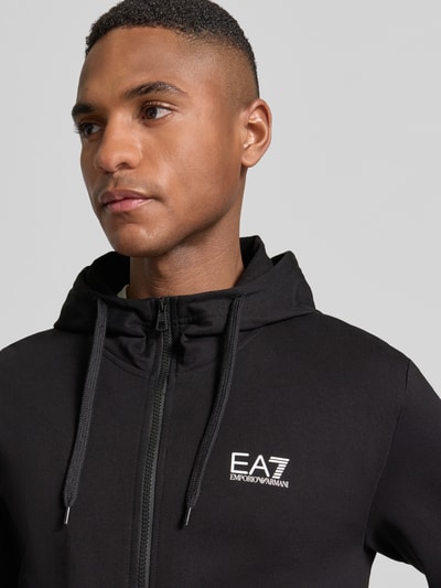 Męskimodel nosi Slim Fit Sweatjacke mit Kapuze od EA7 Emporio Armani, czarny kolor, nadruk z logo, mieszanka bawełny, krój slim fit, zamek błyskawiczny dwustronny, wpuszczane kieszenie, kaptur z tunelem, stójka, elastyczne wykończenia, długość tyłu 69 cm, długość rękawów 69 cm, zbliżenie na górną część ciała
