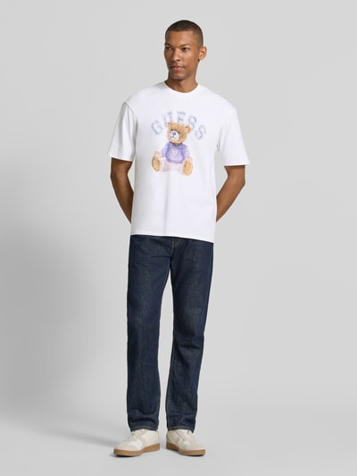 Männermodel trägt T-Shirt mit Motiv-Print und Rundhalsausschnitt von Guess, Herren T-Shirt aus reiner Baumwolle, Regular-Fit, Hauptfarbe Weiß, Motiv mit Bär und Logo, Rückenlänge bei Größe M 68 cm, Ganzkörperansicht von vorne.