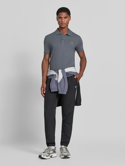 Männermodel trägt Slim Fit Poloshirt aus reiner Baumwolle von Lacoste in Grau, schwarze Hose, neutrale Farben, Ganzkörperansicht von vorne.