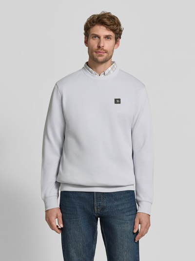 Męski model trägt Sweatshirt mit Label-Patch von Scotch & Soda aus bawełna, luźny krój, okrągły dekolt, prążkowane wykończenia, długość tyłu w rozmiarze M 64 cm, długość rękawa M 64 cm, jasnoszary, kształt prosty, faktura gładka, Ganzkörperansicht von vorne.