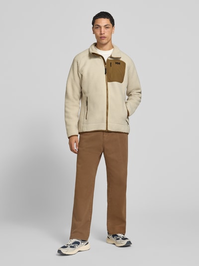 Männermodel trägt Fleecejacke mit Stehkragen von Helly Hansen in Beige mit brauner Napoleontasche, lockere Passform, Ganzkörperansicht von vorne. Rückenlänge 78 cm, Ärmellänge 67 cm, elastische Abschlüsse, seitliche Eingrifftaschen.