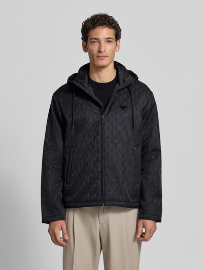 Männermodel trägt Regular Fit Jacke mit Allover-Logo-Print von Emporio Armani, Schwarz mit Kapuze, Zweiwege-Reißverschluss, Ganzkörperansicht von vorne.