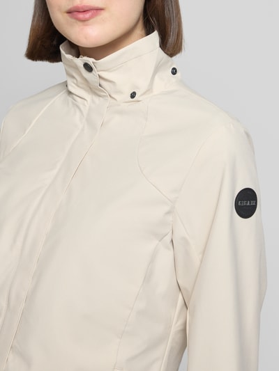 Kobiece modelka nosi Parka mit Kapuze und verdecktem Reißverschluss od G.I.G.A. DX, kremowy kolor, prosty krój, naszywka z logo, detal z zamkiem błyskawicznym, hydrofobowy materiał, długość tyłu w rozmiarze 36: 70 cm, szerokość ramion w rozmiarze 36: 41 cm, szerokość klatki piersiowej w rozmiarze 36: 51 cm, długość rękawów w rozmiarze 36: 63 cm, zbliżenie na górną część tułowia
