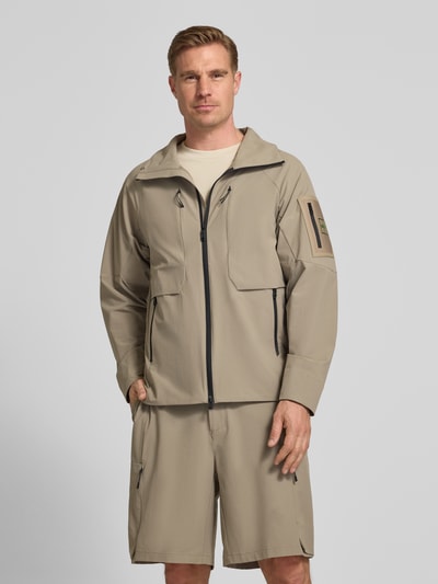 Männermodel trägt Regular Fit Trainingsjacke mit Label Patch Modell 'OW_GLOBE' von BOSS Green in Optik: Unifarben, Zweiwege-Reißverschluss, Stehkragen, Langarm, Reißverschlusstaschen, Ärmeltasche, elastischer Ärmelabschluss; sandfarbene, glatte Textur, Ganzkörperansicht von vorne.