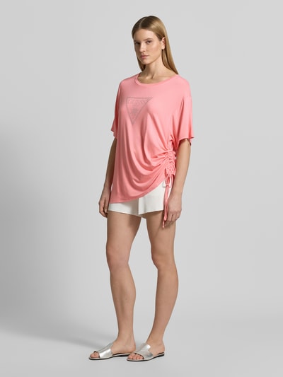 Kobiece model prezentuje T-shirt mit Logo-Print model 'COULISSE' od Guess z mieszanki wiskozy, luźny krój, okrągły dekolt, nadruk z logo, tunel, długość pleców w rozmiarze S: 70 cm, różowy i biały, całkowity widok z przodu