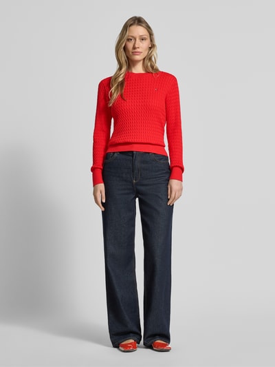 Frauenmodel trägt Slim Fit Pullover aus reiner Baumwolle von Tommy Hilfiger in Rot mit Zopfmuster, Rundhalsausschnitt, langarm, Strickoptik, Logo-Detail, breite gerippte Abschlüsse, kombiniert mit dunkelblauer weiter Jeans, Ganzkörperansicht von vorne.