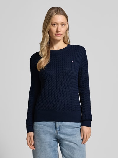 Frauenmodel trägt Slim Fit Pullover aus reiner Baumwolle von Tommy Hilfiger, Optik: Unifarben, Strickoptik, Zopfmuster, Logo-Detail, Ganzkörperansicht von vorne.