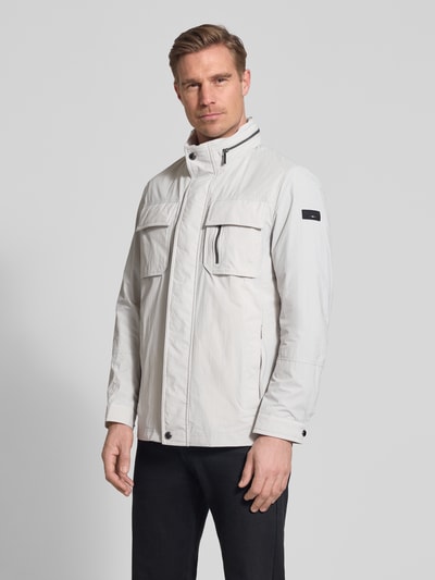 Męski model nosi Regular Fit Jacke mit Stehkragen od Bugatti w jasnoszarym kolorze, z kieszeniami na piersi, aplikacją z logo, regulowanym kapturem, krój regular fit, długość tyłu w rozmiarze 50: 72 cm, szerokość ramion w rozmiarze 50: 46 cm, szerokość w rozmiarze 50: 52 cm, długość rękawów w rozmiarze 50: 68 cm, całkowity widok z przodu.