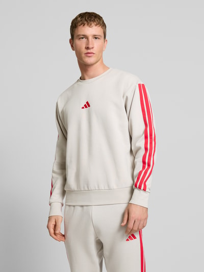 Männermodel trägt Sweatshirt mit Label-Stitching von adidas Sportswear in Beige mit roten Streifen, Regular Fit, Nahaufnahme Oberkörper.