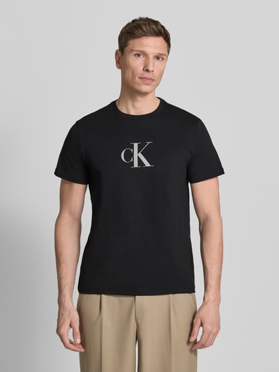 Männermodel trägt T-Shirt mit Rundhalsausschnitt und Logo von CK Calvin Klein in Schwarz, reine Baumwolle, Regular Fit, Ganzkörperansicht von vorne.