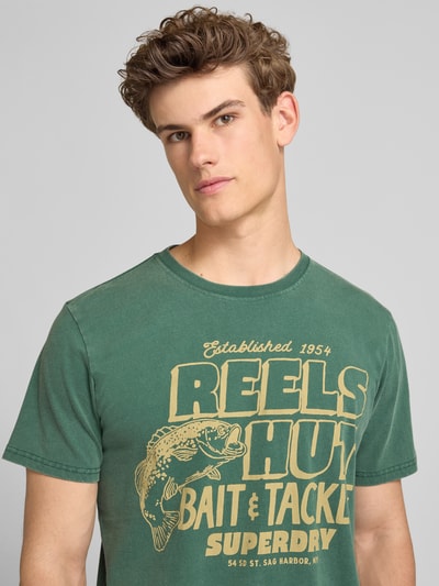 Männermodel trägt grünes Loose-Fit Herren T-Shirt mit Label-Print Modell 'Roadside Trade' von Superdry aus Reine Baumwolle, Rundhalsausschnitt, Nahaufnahme Oberkörper.