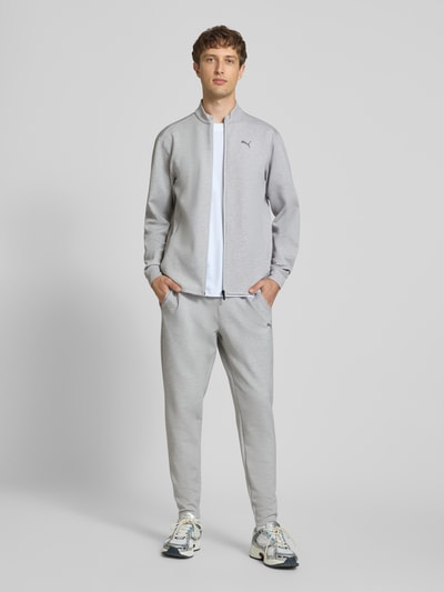 Męski model nosi bluza rozpinana z Mieszanka materiałów z dodatkiem lyocellu, krój regular fit, stójka, logo, zamek błyskawiczny, długość tyłu w rozmiarze M 71 cm, długość rękawów w rozmiarze M 70 cm, Sweatjacke mit Stehkragen und Logo od PUMA PERFORMANCE, jasnoszary kolor, gładka powierzchnia, widoczne kieszenie, całkowity widok z przodu.