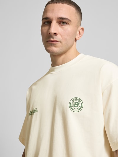 Männermodel trägt T-Shirt aus Baumwolle Modell 'Sunday Miles' von ANOTHER COTTON LAB in Creme mit grünen Label-Details, Nahaufnahme Oberkörper.