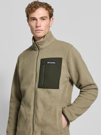 Mężczyzna nosi kurtkę z polaru Columbia Fleecejacke mit Stehkragen Modell 'Jasper' prosta, zamek błyskawiczny, naszywka z logo, kieszeń na piersi, wpuszczane kieszenie, elastyczne zakończenia rękawów, kolor beżowy i oliwkowy, faktura miękka, widok na górną część ciała z przodu.