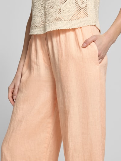 Frauenmodel trägt Leinenhose mit elastischem Bund und Eingrifftaschen von Emily Van den Bergh in Pfirsich, Loose Fit, Nahaufnahme Oberkörper, Reines Leinen, französische Eingrifftaschen, angedeutete Gesäßleistentaschen, Struktur leicht texturiert.
