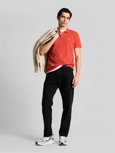 Mannelijk model draagt Regular Fit Poloshirt uit zuiver katoen van Tom Tailor in effen rood, piquétextuur, korte mouwen, logostiksel, polokraag, twee-knoopsluiting, splitjes aan zijkant; vooraanzicht hele lichaam.