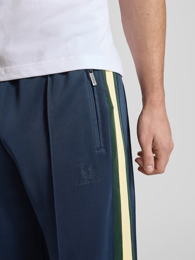 Männermodel trägt Track Pants mit Reißverschlusstaschen und Kordelzug von ANOTHER COTTON LAB in Dunkelblau mit farbigen Seitenstreifen, Nahaufnahme Oberkörper. Material-Mix mit Stretch-Anteil, Wide Fit, elastischer Bund, seitliche Reißverschlusstaschen.