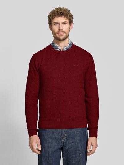 Männermodel trägt Regular Fit Pullover aus Baumwoll-Woll-Mix von GANT, weinrot, dezentes Fischgrätenmuster, Ganzkörperansicht von vorne.
