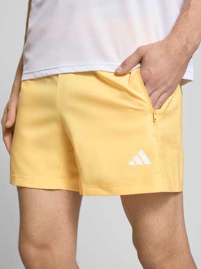 Męski model nosi Regular Fit Sport-Shorts mit Label Print od adidas Training, efekt: jednolity kolor, krój regular fit, nadruk z logo, elastyczny ściągacz z tunelem, kieszenie zapinane na zamek błyskawiczny, Ganzkörperansicht z przodu, detal szortów.