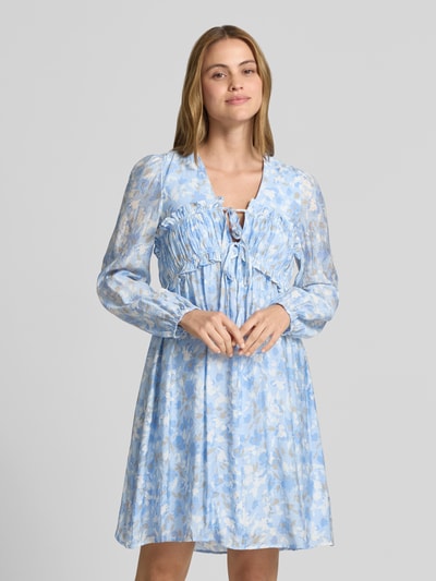 Vrouwenmodel draagt knielange jurk uit viscosemix model 'Pipiana' van mbyM met bloemen all-over print, getailleerde pasvorm, met lange mouwen, ruches, V-hals, elastische mouwboorden; context: dames jurk van mbyM, ruglengte maat S 93 cm, borstwijdte maat S 40 cm, mouwlengte maat S 65 cm. Vooraanzicht bovenlichaam.