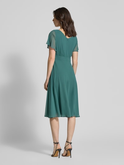Frauenmodel trägt knielanges Kleid mit Rundhalsausschnitt von Vera Mont in Türkis, semitransparentes Design, taillierte Passform, blickdichter Unterrock, Rückenansicht Ganzkörperansicht.