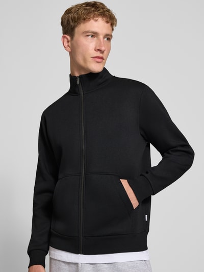 Männermodel trägt schwarze Regular Fit Sweatjacke mit Baumwoll-Anteil Modell 'Bradley' von JACK & JONES, Ganzkörperansicht von vorne. Kontext: Herren Sweatjacke, Material-Mix, Stehkragen, Reißverschluss, geteilte Känguru-Tasche, Rückenlänge 68 cm, Ärmellänge 66 cm.