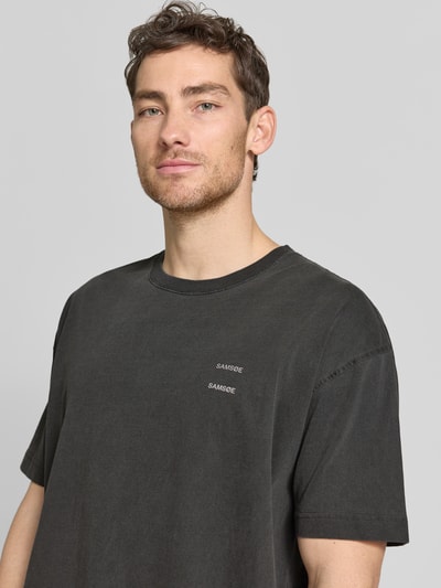 Męski model nosi T-shirt z nadrukiem z logo model 'JOEL' od Samsøe Samsøe, bawełna, krój relaxed fit, okrągły dekolt, długość tyłu 72 cm, rozmiar M, zbliżenie na górną część ciała.
