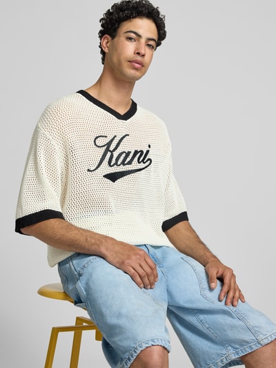 Männermodel trägt T-Shirt in Häkel-Optik mit V-Ausschnitt von KARL KANI, Baumwoll-Mix, Loose-Fit, Label-Stitching, Nahaufnahme Oberkörper.