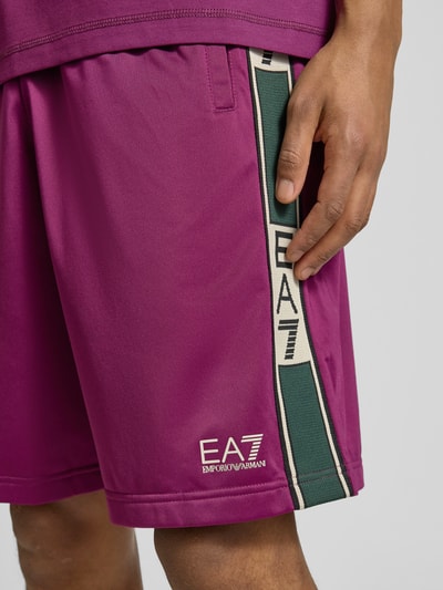 Mężczyzna nosi Szorty mit Logo und Eingrifftaschen von EA7 Emporio Armani w kolorze fioletowym z zielonym paskiem, krój relaxed fit, elastyczny pas, szerokość w rozmiarze M: 42 cm, długość wewnętrzna nogawki: 28 cm, wpuszczane kieszenie, logo, detalowe ujęcie z przodu