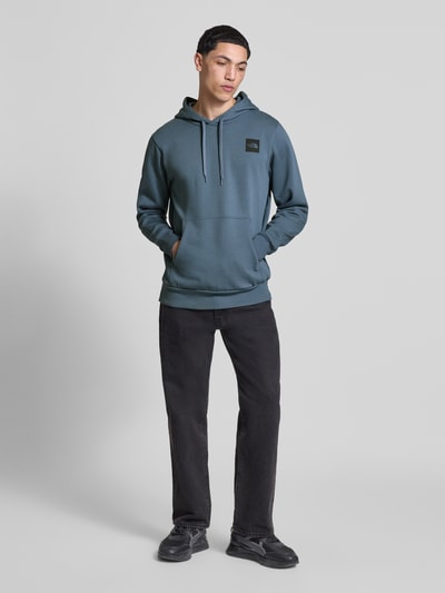 Männermodel trägt Regular Fit Hoodie mit Logo-Print von The North Face in Blau, Kängurutasche, elastische Abschlüsse, Ganzkörperansicht von vorne.