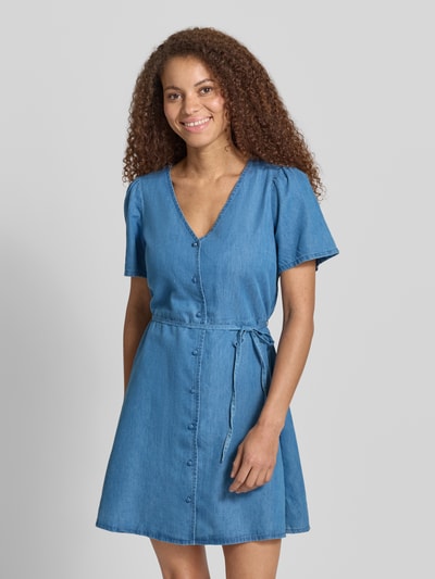 Frauenmodel trägt Tailliertes Jeanskleid aus Baumwoll-Lyocell-Mix Modell 'EVIDA-BEA' von Only, Denim-Optik, Mini-Länge, Knopfleiste, Taillengürtel, V-Ausschnitt, Kurzarm, Ganzkörperansicht von vorne.