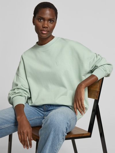 Frauenmodel trägt Oversized Sweatshirt mit Logo-Stitching von Anine Bing in sanftem Mintgrün aus reiner Baumwolle, Oversized-Fit, Rundhalsausschnitt, elastische Abschlüsse, Rückenlänge bei Größe S 63 cm, Ganzkörperansicht von vorne.