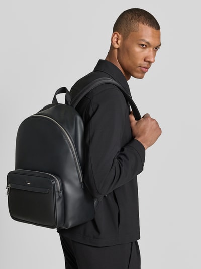 Männermodel trägt Rucksack mit Label-Prägung Modell 'RAY' von BOSS in Optik: Unifarben, Lederoptik, Reißverschluss, Ganzkörperansicht von vorne.