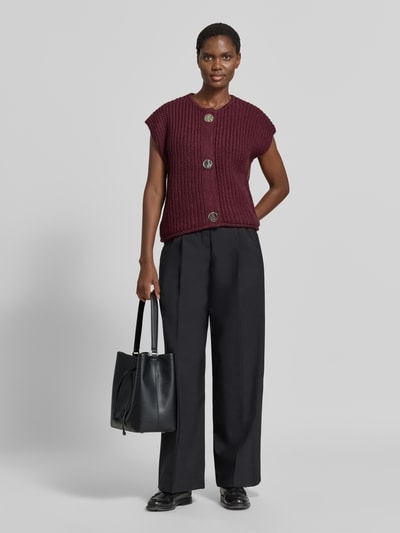 Modelka nosi kamizelkę dzianinową Relaxed Fit Strickweste mit Woll-Anteil Model 'Norma' od b.young, Mieszanka materiałów z dodatkiem wełny, okrągły dekolt, krótkie rękawy, listwa guzikowa, długość tyłu w rozmiarze S: 52 cm, odcień bordowy, spodnie czarne, torba skórzana, zdjęcie typu: całopostaciowe od przodu.