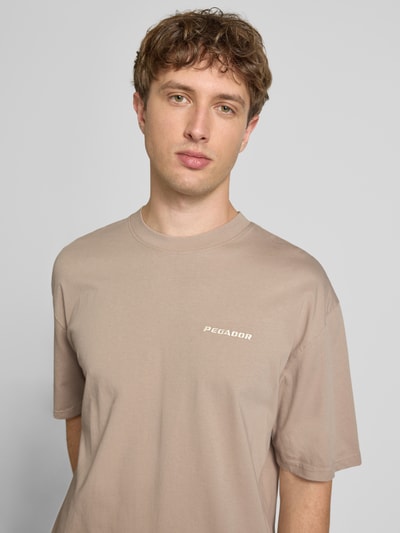 Männermodel trägt Oversized T-Shirt mit Label-Stitching von Pegador aus Baumwoll-Mix in Beige mit Rundhalsausschnitt, Nahaufnahme Oberkörper.