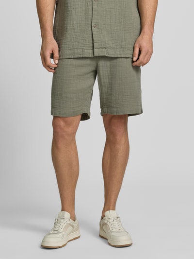 Männermodel trägt Shorts mit Eingrifftaschen und elastischem Bund von Lindbergh in Olivgrün, Reine Baumwolle, Loose-Fit, Nahaufnahme Beine, Ganzkörperansicht von vorne, Bundweite bei Größe M 43 cm, Innenbeinlänge bei Größe M 22 cm.