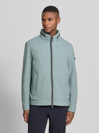 Männermodel trägt Regular Fit Jacke mit Stehkragen von bugatti in Hellblau, Logo-Detail, Zweiwege-Reißverschluss, Eingrifftaschen, Ganzkörperansicht von vorne. Rückenlänge 68 cm, Schulterbreite 42 cm, Brustweite 50 cm, Ärmellänge 65 cm, verstaubare Kapuze, elastische Ärmelabschlüsse.