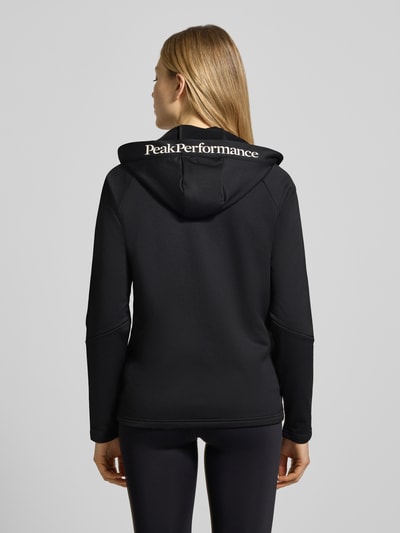 Kobieta nosi Softshell-Jacke mit Kapuze Modell 'RIDER' von PEAK PERFORMANCE, czarna, krój regular fit, tył, materiał elastyczny, detal z logo, całkowity widok z tyłu.