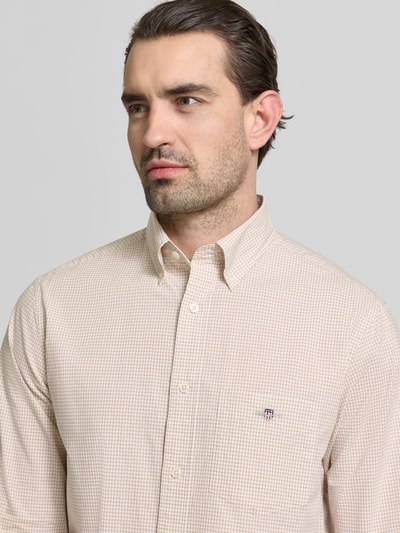 Męski model nosi Slim Fit Freizeithemd mit Logo-Stitching od Gant, beżowa bawełna, krój slim fit, logo, kołnierzyk button-down, długi rękaw, kieszeń na piersi, sportowe mankiety, długość tyłu w rozmiarze M: 79 cm, szerokość ramion w rozmiarze M: 48 cm, długość rękawa w rozmiarze M: 68 cm, gładka tekstura, drobna krata, zbliżenie na górną część ciała, widok z przodu