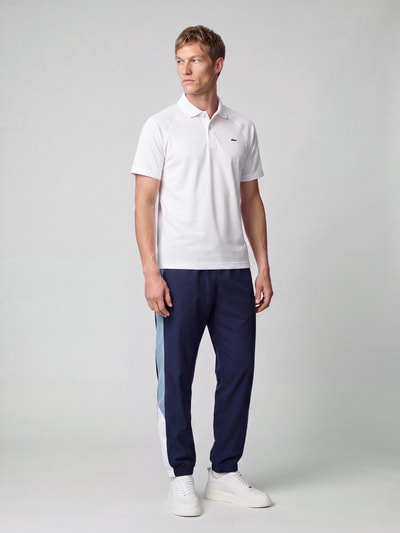 Männermodel trägt weißes Regular Fit Sport-T-Shirt mit Polokragen von Lacoste Sport, dunkelblaue Hose, Ganzkörperansicht von vorne.