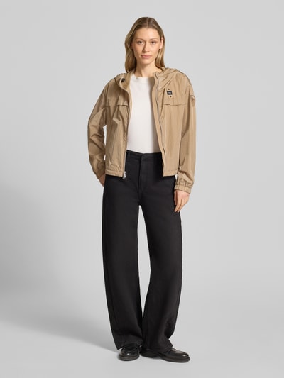 Frauenmodel trägt Trekking Jacke mit Kapuze Modell 'BURRAGE' von Blauer Usa in Beige, gerader Schnitt, zweiwege Reißverschluss, seitliche Reißverschlusstaschen, elastische Ärmelabschlüsse, Label-Patches auf Brust und Ärmel, glatte Textur, kombiniert mit schwarzer Hose und weißen Schuhen, Ganzkörperansicht von vorne.