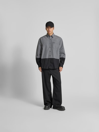 Männermodel trägt Relaxed Fit Freizeithemd mit Brusttasche von VETEMENTS im Baumwoll-Mix, zweifarbig, durchgehende Knopfleiste, Langärmlig, Ganzkörperansicht von vorne.